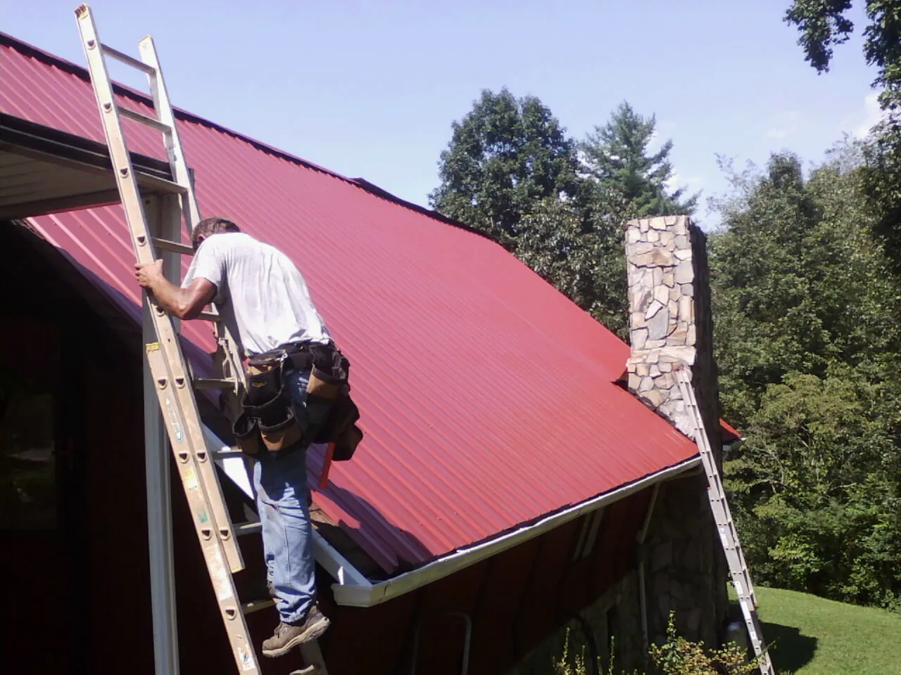 Roofer on a ladder beside a red metal roof and stone chimney — Soffit & Fascia Repair by Roofing Brothers in Sadsburyville
